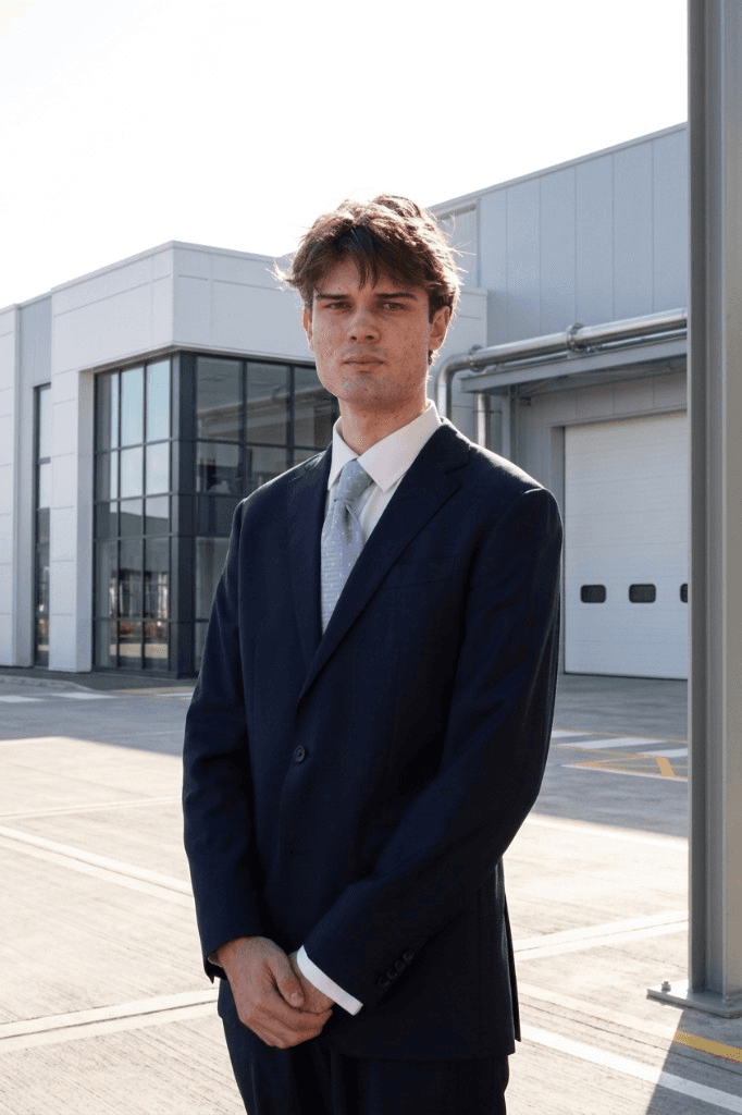 Jonty MacIntyre standing outside an industrial building in New Zealand, wearing a navy suit and light blue tie