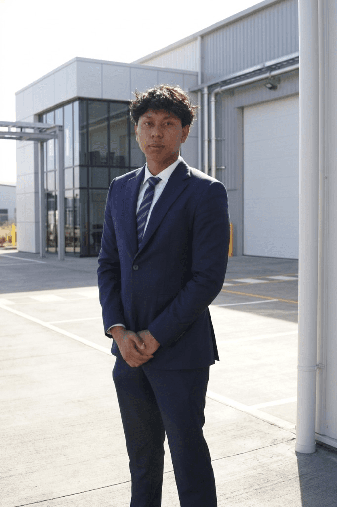 Zak Rachmadi standing outside an industrial building in New Zealand, wearing a navy suit and striped tie